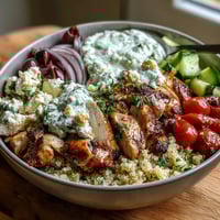 Freshly grilled lemon-herb chicken slices rest over fluffy quinoa in a Greek Chicken Power Bowl, topped with crisp cucumber, cherry tomatoes, red onion, and creamy tzatziki.